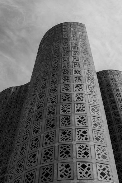 A black and white photograph of the tall, curved, undulating structure containing the Great Hall in the New York Hall of Science museum, originally the 1964 World Fair science pavilion. The photo was taken at the base of one of the curved sections, looking upward. The section looms, tower-like. The adjacent section of wall is behind to the left partially in shadow. To the right, the wall of the hall itself is just visible behind the foreground section. The entire surface of all the walls are covered with rectangular panels each embedded with irregular, dark glass shapes that serve to let some light into the interior.