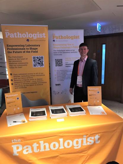 A photograph of The Pathologist's conference booth, displaying print copies of the magazine, registration forms, and business cards. Business Development Executive Will Whittaker stands in the background.