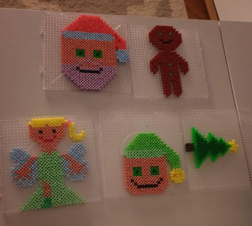 Auf einem weißen Tisch sind 5 quadr. Bügelperlenvorlagen. Darauf ist oben links ein Weihnachtsmann mit lavendelfarbenem Bart und hellblauem "Fell" an der Mütze, daben ein Lebkuchenmann mit grünen Augen u Knöpfen. 
Unten v.l.n.r.: Engel mit blondem Pferdeschwanz, grünem Kleid und blauen Flügeln, dann Elfengesicht mit grüner Mütze, dann ein Tannenbaum mit Sternspitze.