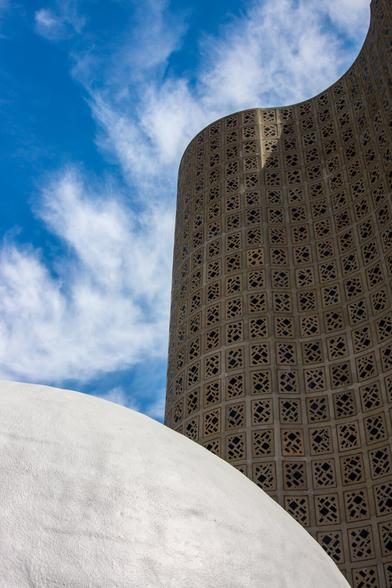 A color photograph of the outside of the New York Hall of Science museum, originally the 1964 World’s Fair science pavilion. The curved, tan, concrete wall  of the Great Hall rises on the right, covering about half the frame a bit in the background, pockmarked with its oddly shaped, dark glass windows embedded in rectangular panels matching the wall in color.  At the bottom of the frame in the foreground, a white dome, cropped at its highest point by the left edge of the photograph, slopes to the right ending just before the right edge of the frame. Puffy white clouds and a bright blue sky are visible.