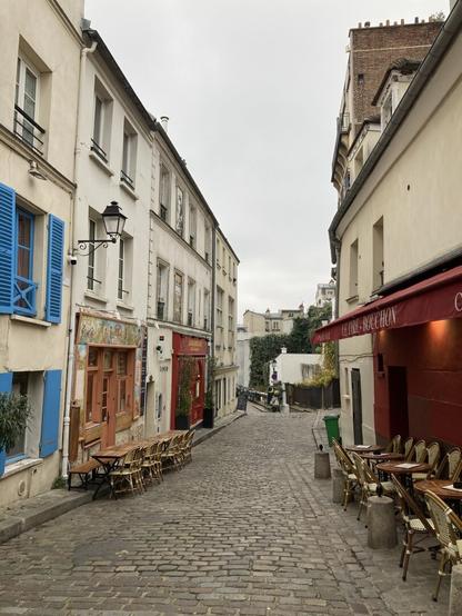 a mostly monochrome street in the Montmartre neighborhood of Paris, except for a set of bright blue shingled windows, one resto and one cafe with red entrances, another with decorative tiles.  Empty cafe tables and chairs sit outside waiting for the lunch rush. 2 sandwich signs - 1 with a restaurant menu, 1 with a drawing of a singer playing a sitar - sit at the end of the cobblestone street as it divides into 2 directions, enticing potential customers.