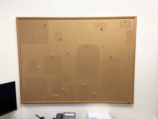 A large empty bulletin board shows shadows where things were once pinned to it. The shadows are darker because for years the items pinned protected the cork from light and oxidation. There are pins of various colors and styles scattered about the board roughly where they were used to affix items