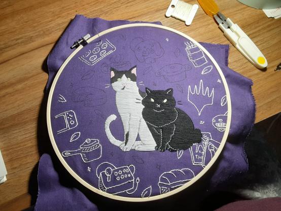 A partially finished embroidery piece showing two stylized cats and the outlines of lots of different objects surrounding them