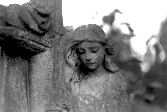 A black and white film photograph of a detail of a gravestone in the form of a female figure clinging to a cross.