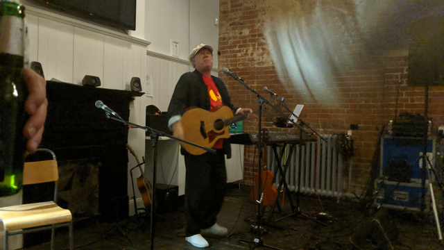 Neil Innes in Ipswich. Neil is wearing a red t-shirt, black shirt and trousers and a white cap. He is holding his guitar and is standing next to his keyboard. He is surrounded by speakers, cables and other instruments.