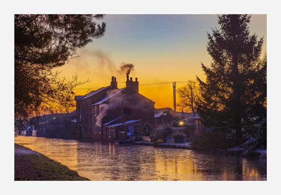 Just before sunrise, looking across a frozen Canal at a house that is silhouetted against the pre dawn glow. The house is coming to life with the chimney showing the first fire of the day. Cold, frosty outlook.
