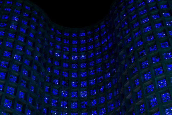 A color photograph, angled up at the interior wall of the darkened Great Hall in the New York Hall of Science museum, the former 1964 Worlds Fair science pavilion. The curved, ribbonlike walls are, from floor to ceiling, largely comprised of concrete panels with translucent blue glass shapes embedded in them which glow from the sunlight outside, giving it an otherworldly feeling. The walls vanish into the black ceiling.
