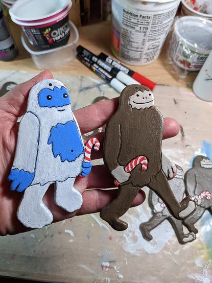Two whimsical ornaments made of clay and acrylic, depicting a yeti and Bigfoot, smiling and carrying candy canes.