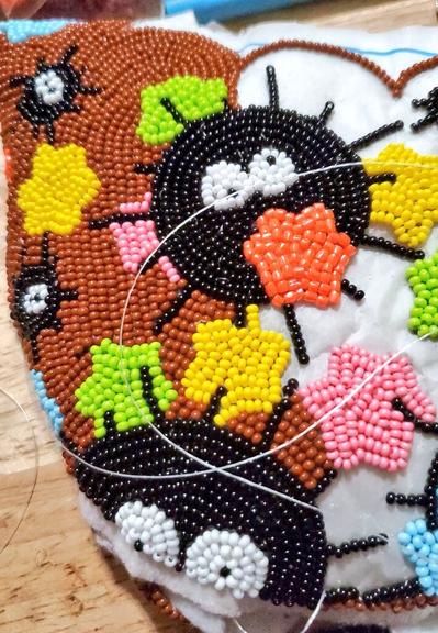 Work in progress of beaded soot sprite moccasins. Brown background being filled in. Soot sprites are black circles with wisker like spikes around them and googly eyes. Little stars are in colors of yellow, pink, green, orange, and blue.