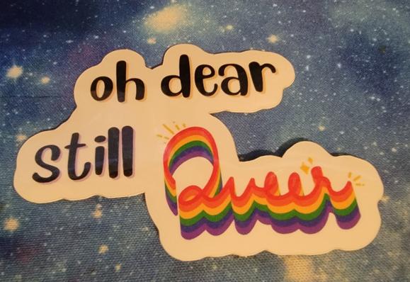 Sticker reading "Oh dear still queer" with queer written in a rainbow shaded cursive on a galaxy mat background.