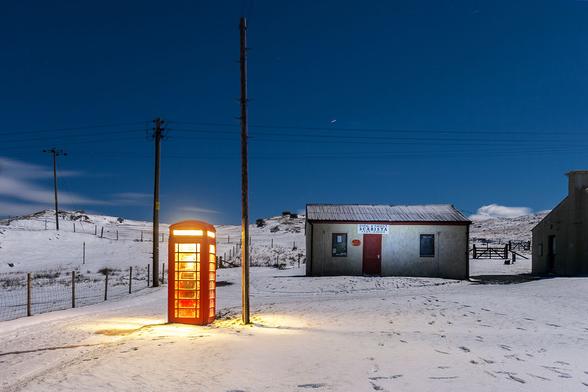 Red telephone box in the snow, lit up at night, next to a small post office building.