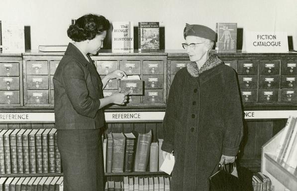 A young Filipino woman is opening a card catalogue while an elderly woman in glasses watches. The photo is black and white, and there are several reference books on shelves around the two individuals.