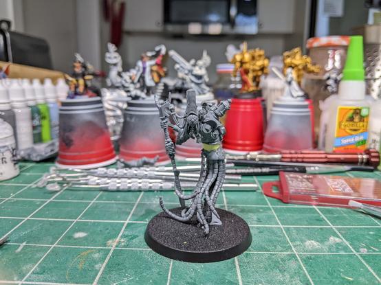 A sinister figure whose legs have been replaced with a dozen mechanical tendrils and leaning on a techno-arcane scepter. Their face is obscured by an insect-like visor covered in many lenses.