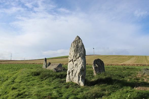 Blue sky with quite a lot of high pale cloud. The sun is shining, it's late afternoon, autumn. A harvested field leads up to a wind turbine. The circle stands in an area of rough grass in a field of... some sort of low-growing green crop. A tall, pointed standing stone at the front, and lower stones behind including a 3.8m recumbent with a pointy flanker. Originally twelve stones, four standing, four falling, four missing.