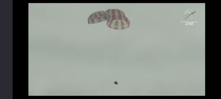Screen capture of the Artemis I Orion capsule descending under parachutes taken from the NASA YouTube live broadcast.. The capsule appears as a dark truncated cone at the bottom of the frame. Above it are the three main parachutes, fully inflated. The parachutes appear as semicircles with vertical orange and white stripes. The sky in the background appears a light grey.

A NASA logo watermark is in the top right of the frame.