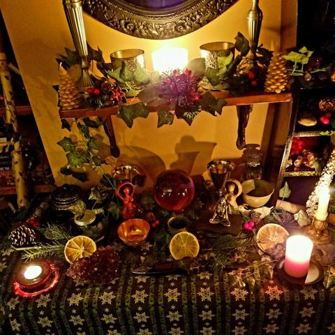 Yule altar with candle sticks and candles in holders, tree shaped beeswax candles, greenery, goddess statues, black and gold incense burner, copper offering bowl, lavender crystal ball, Gaia statue, amethyst cluster, silver and pottery chalices, tiny goat statue, spell bottles,  dried orange slices, cone and berry sprigs, sgian-dubh in a black and silver tipped sheath with knot work, all sitting on 2 shelves with a green table cloth on the lower one with white snowflakes and scroll work.