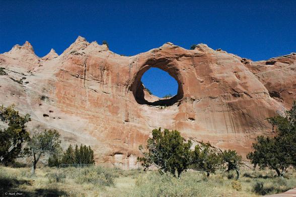 An ochre coloured sandstone rock face with a circular porthole window that has been eroded through the middle of it. The deep blue sky is visible through the porthole and above the rock face. In the foreground are some scrubby trees and long dry grass.