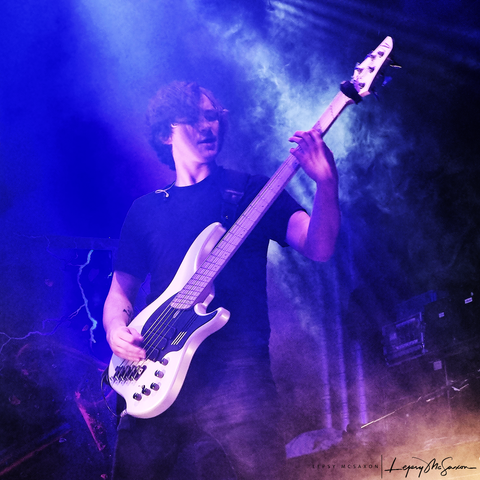 Dominik Gusch steht auf einer in blaues Licht getauchten Bühne und spielt konzentriert Bass. Um ihn herum wabern Nebelschwaden.
Dominik Gusch is standing on a stage doused in blue lights and is immersed in his bass play. He's surrounded by whisps of fog.