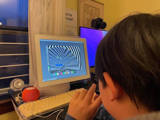 An over-the-shoulder shot of a kid playing with Broderbund's Kid Pix on an iMac G4. The drawing is a checkerboard spiral pattern with sprites on it.