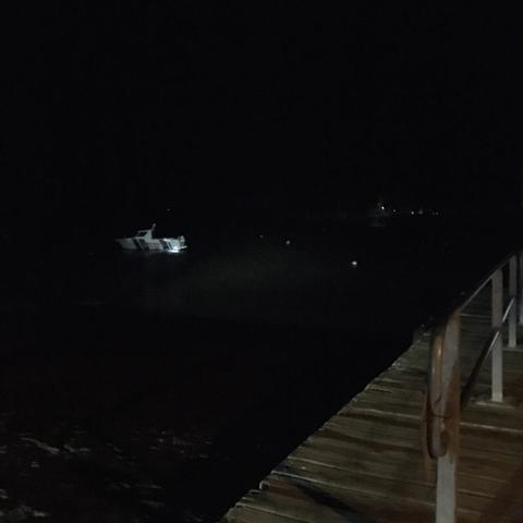 A photo taken in the dark overlooking water from the end of a wooden jetty, there is a boat in the water, there are lights the other side of the estuary. Waves are gently lapping the shore.