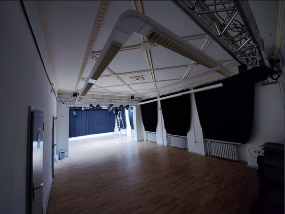 Ein kliner Saal, mit abgehängtem Licht und Parket. schwarze Vorhänge in den Fensternischen, im Bühnenraum ist es hell, eine Trittleiter steht, der Raum der Zusehenden bei der Kamera ist leer und dunkel.