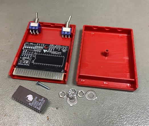 Commodore 64 cartridge parts on my desk. A red iComp cartridge case, a Versa64 cart PCB, a 27C256 EPROM and two switches. The components are placed in the cartridge case for checking the dimensions.