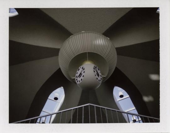 A view from below of a spherical clock hanging from a circular support in a ceiling where the white, curved, vaulted elements converge. A handrail for a walkway is visible at the bottom, and two oblong windows are in the ceiling behind, symmetrical on either side of the clock.