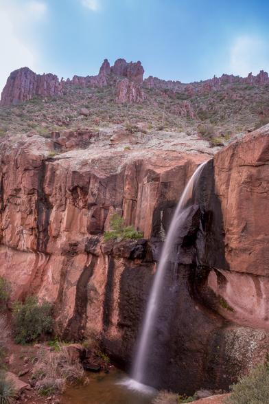 A small cascade of water drops from a tall height over a sharp cliff of red rock. The rock behind the small waterfall is dark and wet while the surrounding rock is dry and dusty. Above the falls, there are some green shrubs and cacti on a gentle slope up to some more jagged rock outcrops (I'm not sure if they're called hoodoos down here, but they look a bit similar to them, again in a redish rock). The sky above is blue with a few light clouds and some sunflare.