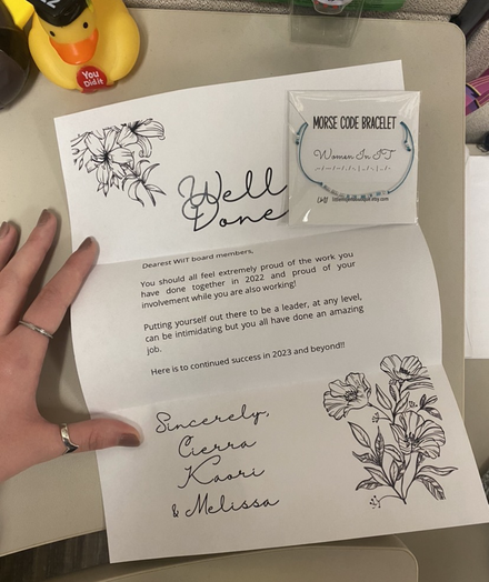 A white letter with flowers in the corner stating: “Well Done. Dearest Wit board members, you should all feel extremely proud of the work you have done together in 2022 and proud of your involvement while also working! Putting yourself out there to be a leader, at any level, can be intimidating, but you all have done an amazing job. Here is to continued success in 2023 and beyond! Sincerely, Ciarra, Kaodi, and Melissa”. Included in the top right corner is a blue bracelet that says “Women in IT” in Morse code.
