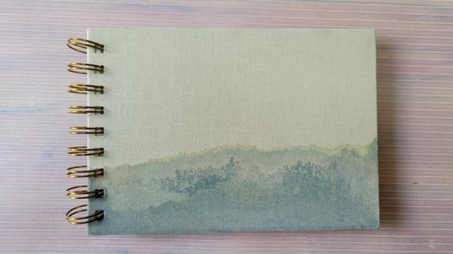 Small horizontal sketchbook with light taupe linen cover and large brass double wire rings - the upper third of which is blank and the lower third of which is taken up with what appears to be a mountain range in tonal variations of grayish-greens - somewhat like a view of the rolling Blue Ridge Mountains of the southeastern U.S.