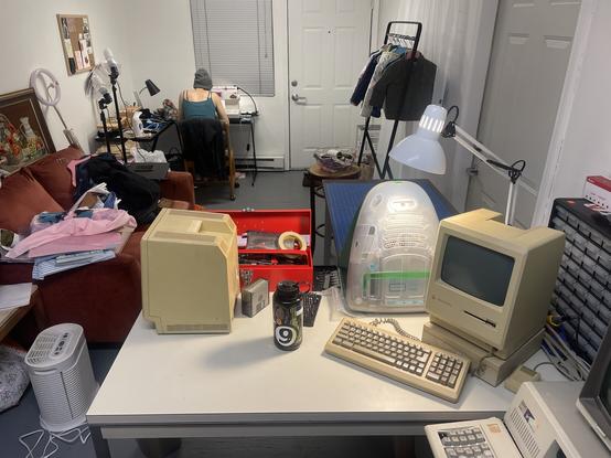 Photo of an art/work studio. In the foreground are several vintag Macintosh computers and a toolbox, in the background a woman sits at a sewing table.