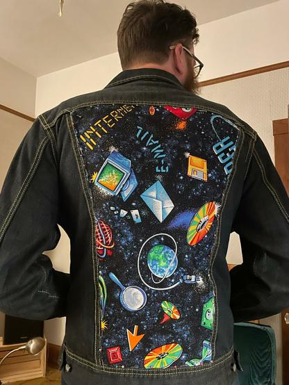 The back of a man standing in a denim jacket with a patch bearing brightly colored 90's era internet icons and words such as 'Internet' and 'E-Mail' on a space-themed background.