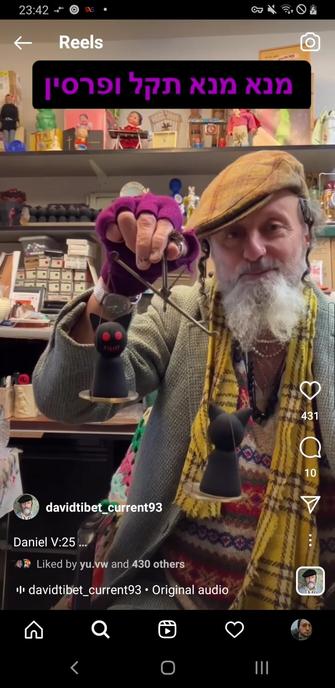 Still frame from musician David Tibet's (of Current 93 fame) instagram is depicting him as eccentricly-dress man with a white beard and a flat cap. He handles simple scales with a clay red-eyed demon on the each side. Caption above Davit states in Hebrew: mene mene tekel uparsin.
