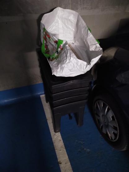 Bac de lombricompist noir installé sur la place de parking a coté d'une clio dans un sous sol d'immeuble un sac blanc carrefour fixé a une planche a roulette DiY est posé dessus (on comprend qu'elle a servi au transport dans l'ascenseur)