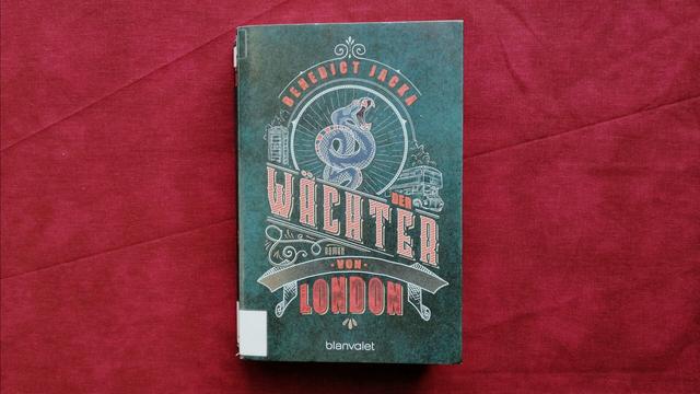 The German edition of part 4 of Benedict Jacka's Alex Verus series lies on red fabric. The title reads "Der Wächter von London" (translates to "The Guardian of London", original title is "Chosen").