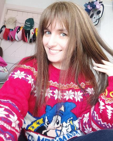 Selfie of myself with a new hair cut. Hair is straight and below shoulder length, and I have a fringe. Blonde highlights are scattered through my dark brown natural colour. I'm wearing a red Sonic the Hedgehog Christmas jumper.