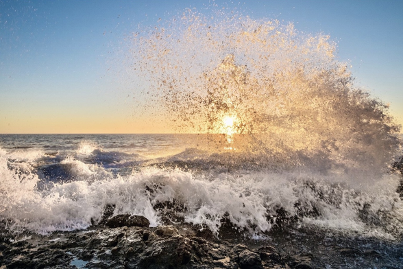 Solar flare. A wave crashing into the rocky coastline projects a circular splash in-line with the sun setting giving the impression of a solar flare.