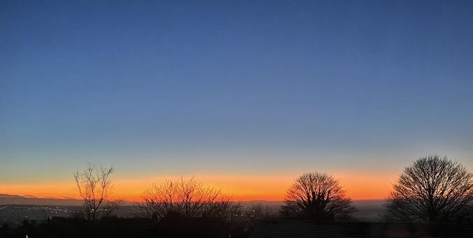 A photograph of the twilight, looking out over the Cheshire Plain. The top of the photo is blue, fading to paler blue as the sky dips to the horizon where it meets a band of bright orange. There are two trees in the foreground and the distant lights of Kidsgrove off to the left.