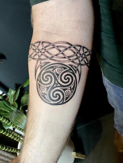 A man’s arm with a large black and gray tattoo of a Triskelion inside a circle, decorated with Celtic patterns, attached to a band of knotwork encircling the arm, rendered in fine lines and dot work.
