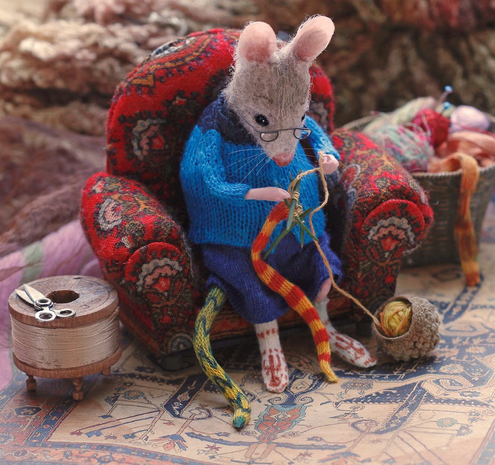 A handcrafted miniature scene with a mouse or rat sitting in a red paisley easy chair surrounded by miniature sewing and knitting notions. The mouse or rat is wearing a knitted peacock blue sweater with a navy blue yoke, and matching navy blue knitted skirt and knitted red and white patterned socks and a knitted yellow and green "tail sock." It's wearing spectacles and knitting an orange and yellow striped "tail sock."