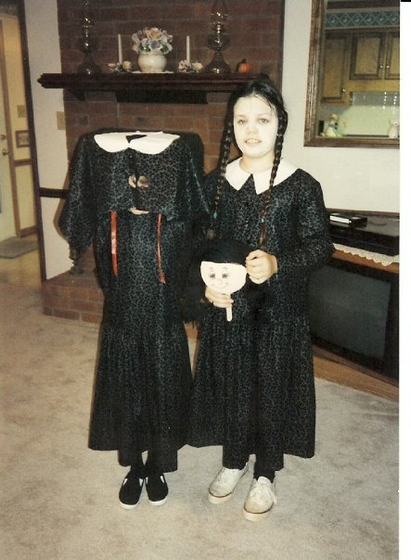 two young girls standing in a 90s living room. The girl on the right is dressed as Wednesday Addams (all black, pigtails, pale face) and holding a doll head on a stick. The girl on the left is wearing an identical dress but is wearing a box over her head to simulate that she has no head.