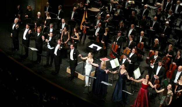 Les chanteurs et chanteuses (Sabine Devieilhe en robe blanche au centre) se tiennent par la main pour les saluts, devant l'orchestre debout.