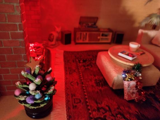 A photograph of a dolls house livingroom that is bathed in a warm orange glow of firelight from the hearth. At the front left is a small decorated Christmas tree with a star on top. Leaning against a footstool is a gift bag with a picture of Santa on it. The bag is filled with tinsel which is spilling onto the floor. On a tray on the footstool is a box of Christmas baubles, a cup of tea and a bar of chocolate. On the far side of the hearth a cat sits on a mat and eyes up the tree. The corner of a sofa can just be seen to the right. It looks like a cosy and relaxing place to sit.
