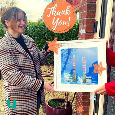 Deborah is presenting a framed print of the Poolbeg Chimneys, a Dublin landmark, to an unseen person in a doorway.