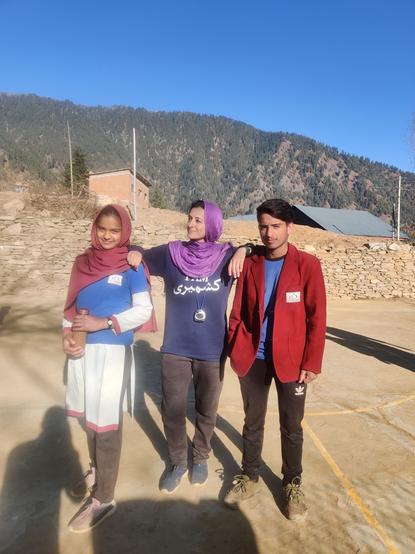 Lady in black tracks and blue shirt, with purple hijab, standing with w her elbows resting on either side, on students: screen right, one dark haired teenage boy in a maroon school blazer, tracks and standing casually with hand in pocket; screen right, teenage girl in school uniform and tracks,with maroon hijab, holding a bottle.