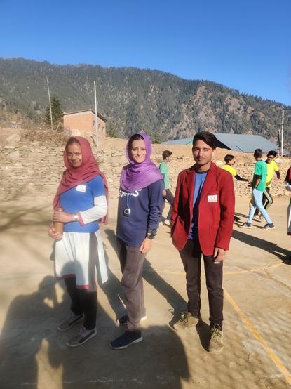 The people looking into camera, sun shining on them. Center: Lady in black tracks and navy blue shirt, with purple hijab, posing sideways. Standing on either side, students: screen right, one dark haired teenage boy in a maroon school blazer, tracks and standing casually with hand in pocket; screen right, teenage girl in school uniform and tracks,with maroon hijab, holding a bottle.