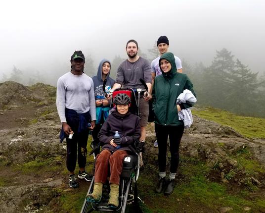 Me (sitting in trail rider) & Power To Be adaptive recreation staff & volunteers, atop Little Mount Doug summit.  Foggy background.
