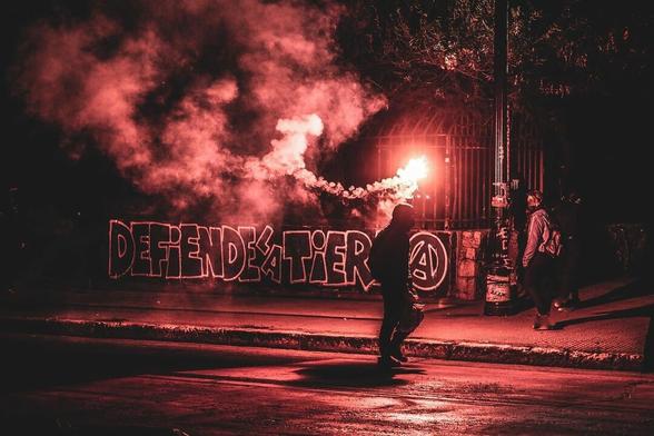 A Chilean anarchist holds a burning red flare in front of a graffiti mural reading "Defend the Earth" in Spanish with a circle-a.