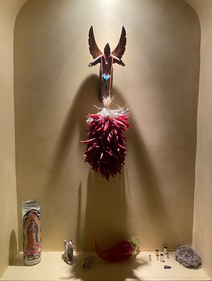 A nicho displaying a santo, a ristra, a Virgin of Guadalupe candle, a small sculpture of a dog with wings, a hummingbird nest, and various found objects