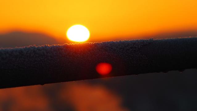 Eis auf einem Handlauf. Im Hintergrund verschwommen  die Sonne, welche alles in ein orangenes Licht taucht